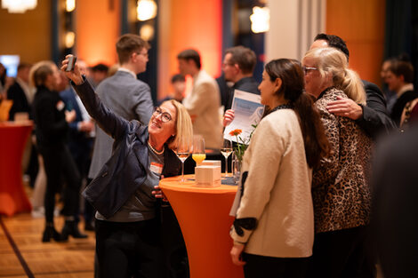 Mehrere festlich gekleidete Personen stehen an einem Stehtisch, eine von ihnen macht ein Selfie von sich und den anderen. Im Hintergrund sind weitere Menschen zu sehen.