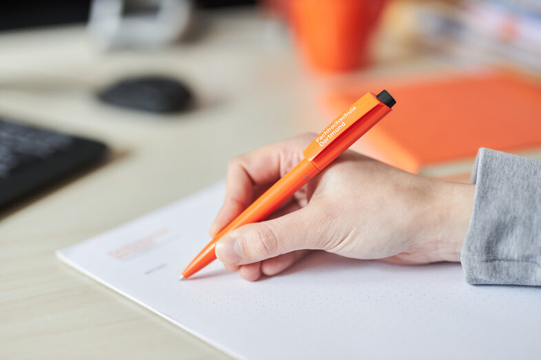 Photo of a hand writing on a notepad with a ballpoint pen. The pen is orange and bears the University of Applied Sciences lettering in white.