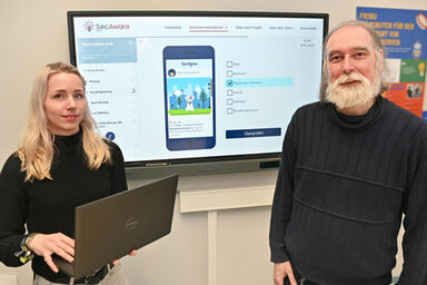 Zwei Personen stehen vor einem großen Bildschirm, auf dem die digitale Lernplattform "SecAware".nrw zu sehen ist.