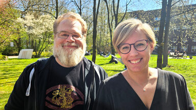 Two people stand in the sunshine in a green courtyard and look friendly into the camera.