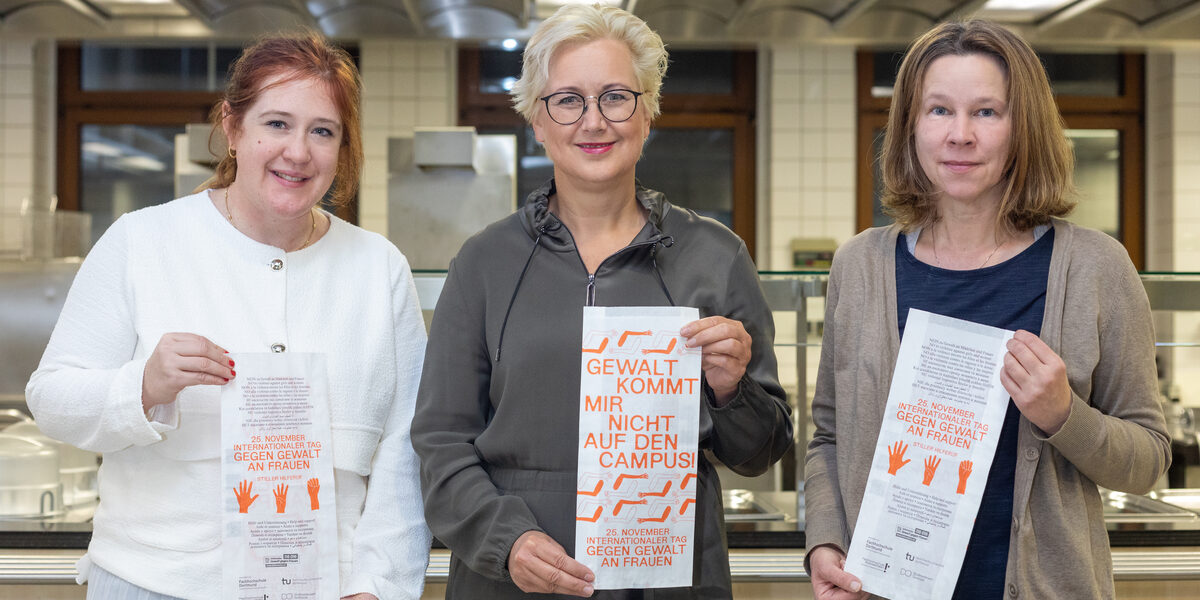 Drei Frauen stehen nebeneinander vor einer Großküche. Jede von ihnen hält eine Papiertüte vor sich.