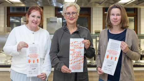 Three women stand next to each other in front of a canteen kitchen. Each of them is holding a paper bag in front of her.