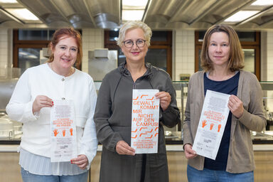 Drei Frauen stehen nebeneinander vor einer Großküche. Jede von ihnen hält eine Papiertüte vor sich.