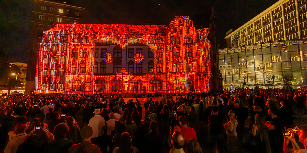 One building is completely illuminated in orange, with the letters DO in the middle. Countless people are standing in front of it in the dark.