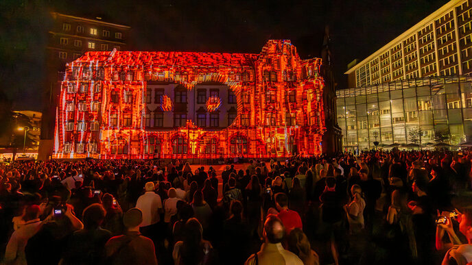 One building is completely illuminated in orange, with the letters DO in the middle. Countless people are standing in front of it in the dark.