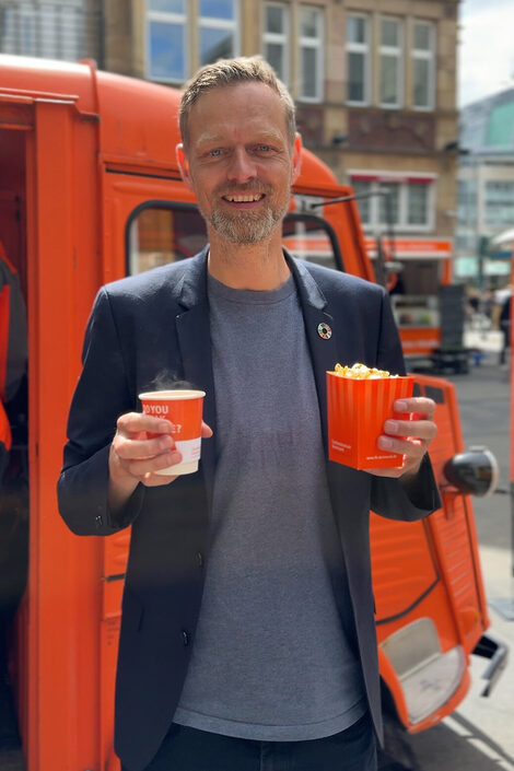 A person stands in front of a classic car with a paper cup and a packet of popcorn.