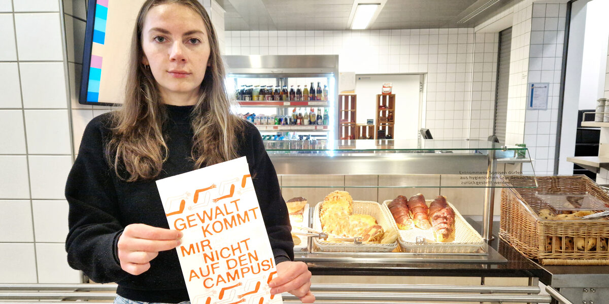 A person stands in front of a counter with baked goods in the canteen and holds a bag in front of them with the inscription: "Violence doesn't come on campus!".