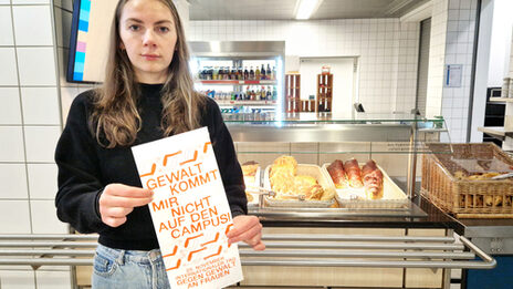 A person stands in front of a counter with baked goods in the canteen and holds a bag in front of them with the inscription: "Violence doesn't come on campus!".
