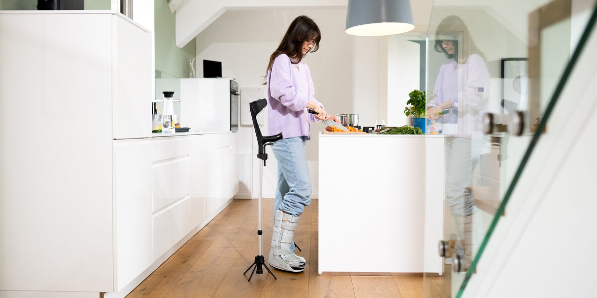 Eine weiblich gelesene Person steht in einer Küche. Sie hat den rechten Fuß ein einer Schiene. Neben ihr steht die Gehhilfe mit dem STEETS-Modul.