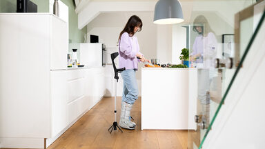 A person read as female is standing in a kitchen. She has her right foot in a splint. Next to her is the walking aid with the STEETS module.