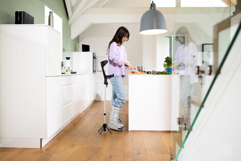 A person read as female is standing in a kitchen. She has her right foot in a splint. Next to her is the walking aid with the STEETS module.