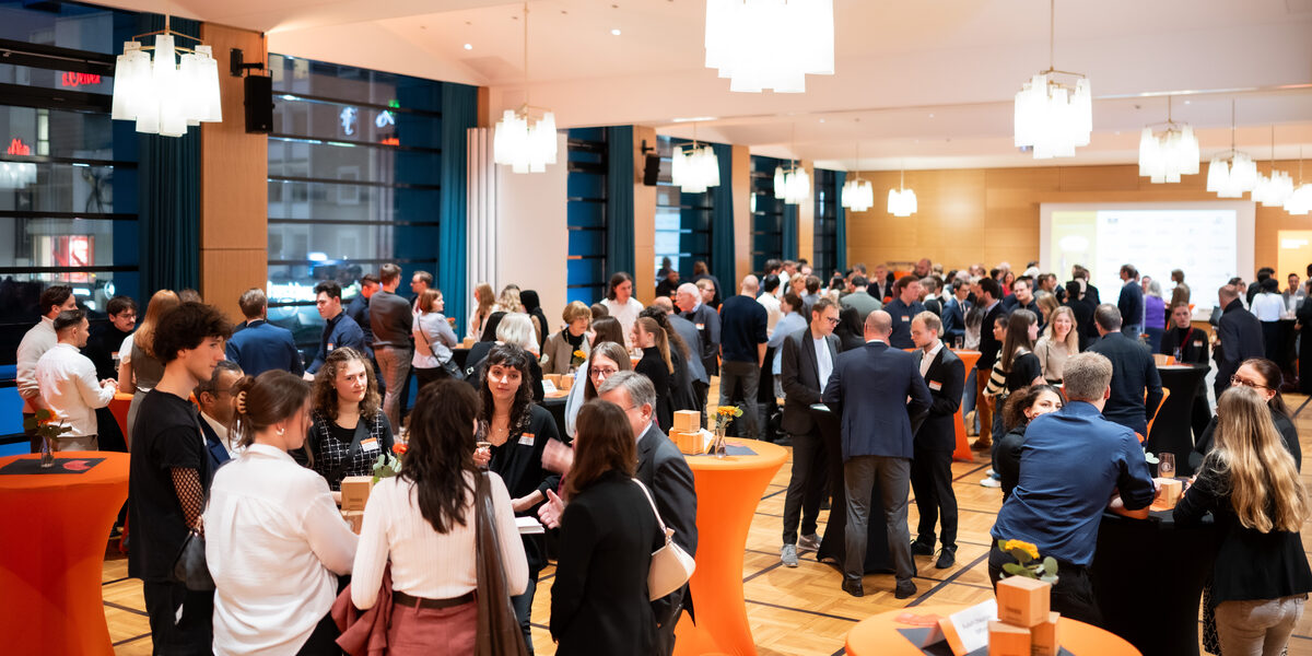One room is festively lit with ceiling lamps, several people stand in groups at tables and chat.