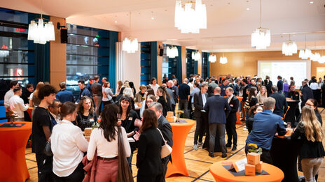 One room is festively lit with ceiling lamps, several people stand in groups at tables and chat.