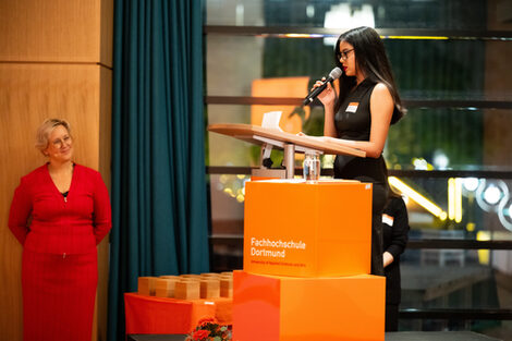 A person stands at a lectern with a microphone in their hand. Two large orange cubes are set up in front of her and a table with several smaller wooden cubes is placed next to her. One person stands at the edge and smiles at the other person.