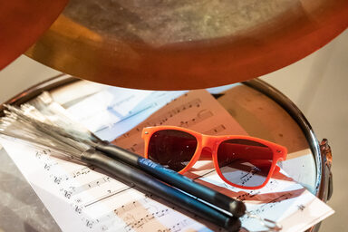 Photo of a drum kit. On it are a sheet of music, a pair of sunglasses and two drumsticks.