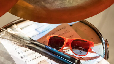 Photo of a drum kit. On it are a sheet of music, a pair of sunglasses and two drumsticks.