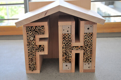 Wooden insect hotel in the shape of a house with the letters "F" and "H", filled with tubes and drill holes as nesting sites.