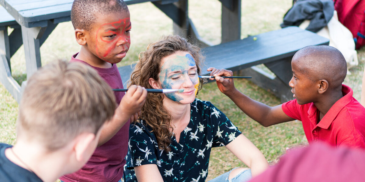 Eine Studierende wird von Kindern in Südafrika mit Pinseln im Gesicht bemalt.