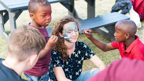 Eine Studierende wird von Kindern in Südafrika mit Pinseln im Gesicht bemalt.