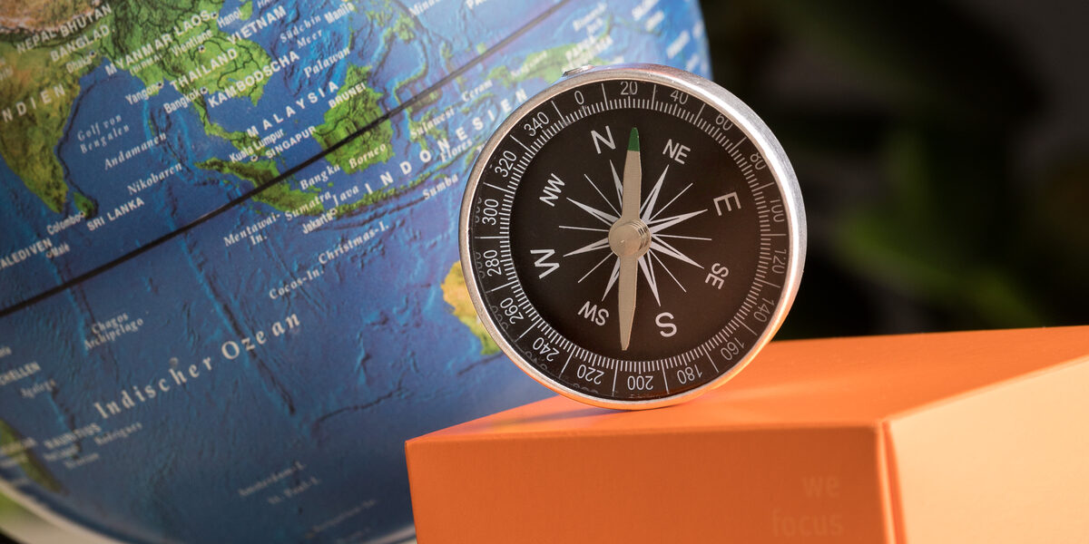Photo of a still life: globe in the background, FH cube, orange paper airplane and compass in front of it.