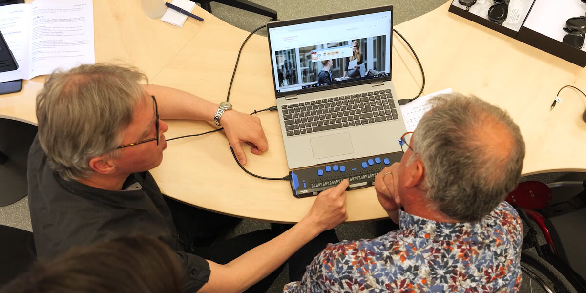 Two people are sitting at a table. And they are looking at a laptop. One person shows the other a device that blind people can use to capture the contents of the screen.
