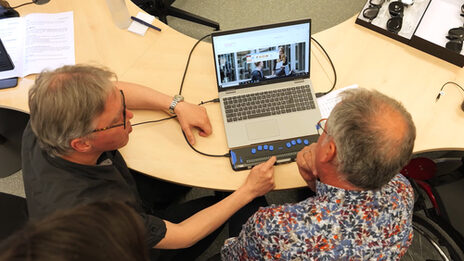 Two people are sitting at a table. And they are looking at a laptop. One person shows the other a device that blind people can use to capture the contents of the screen.