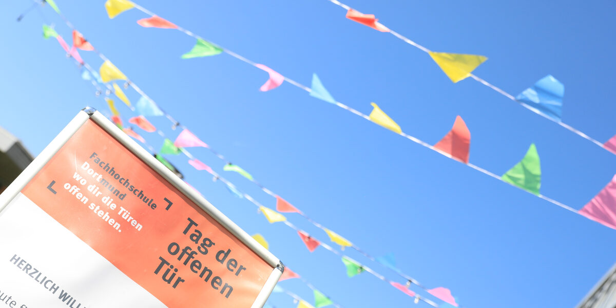 Vor blauem Himmel flattern farbige Wimpelbänder. Links vorn ragt ein Plakat mit der Aufschrift „Tag der offenen Tür“ ins Bild.