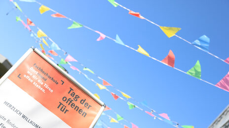 Vor blauem Himmel flattern farbige Wimpelbänder. Links vorn ragt ein Plakat mit der Aufschrift „Tag der offenen Tür“ ins Bild.