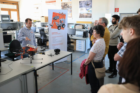 People in front of a lab bench, behind which a person speaks and points to a drone on the bench.