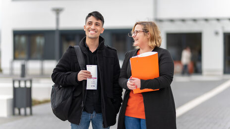 Foto von einer Mitarbeiterin und einem Schüler, die vor einem Campusgebäude stehen. Sie hält eine orangene Mappe unterm Arm, er hat einen Flyer in der Hand. Beide lachen.