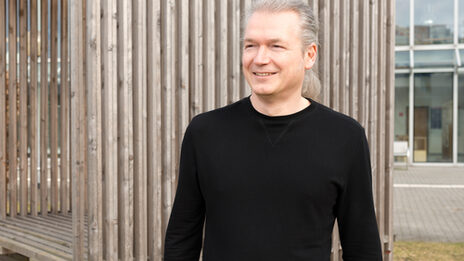 A person stands smiling in front of a wooden construction and looks to the side.