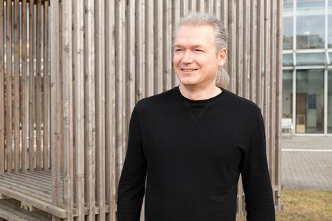 A person stands smiling in front of a wooden construction and looks to the side.