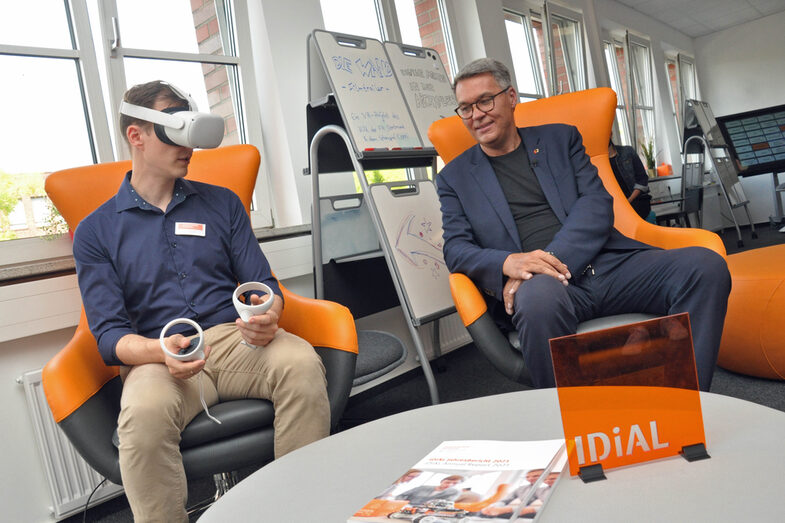 Zwei Personen sitzen auf zwei Sesseln nebeneinander. Der FH-Mitarbeiter links hat eine VR-Brille aufgesetzt und hält zwei Steuerungselemente, der Oberbürgermeister schaut zu.