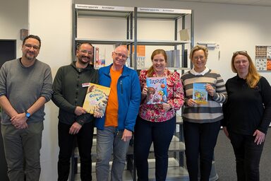 Sechs Personen mit einigen Kinderbüchern in den Händen stehen in einem Raum und schauen in die Kamera.