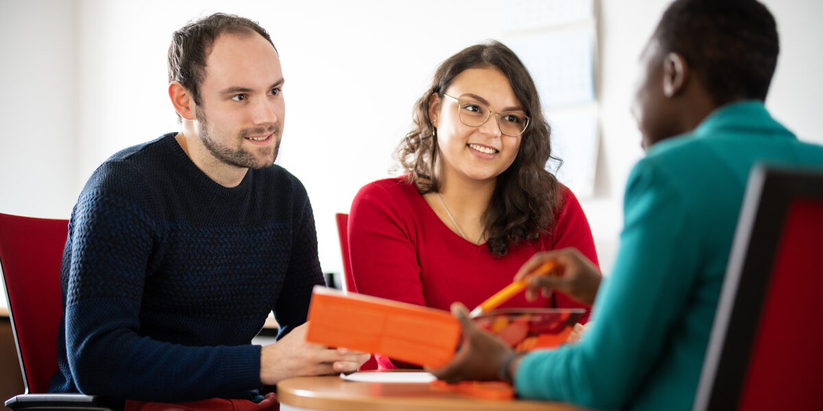 Photo of an employee advising two students sitting opposite her at a table. She points to the flyer she is holding in her hand. The students are in focus,