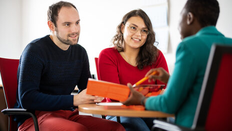 Photo of an employee advising two students sitting opposite her at a table. She points to the flyer she is holding in her hand. The students are in focus,