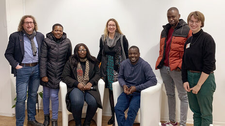 Seven people, some standing, some sitting, in front of a white wall look into the camera.