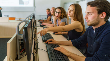 Foto von fünf Studierenden in einer Reihe an PCs. Eine Studentin zeigt den anderen etwas auf dem Bildschirm ihres Sitznachbars.__Reihe an fünf PCs und schauen auf die Bildschirme.__Five students sit in a row at five PCs. One student is showing something to the others on the screen of the student sitting next to her.