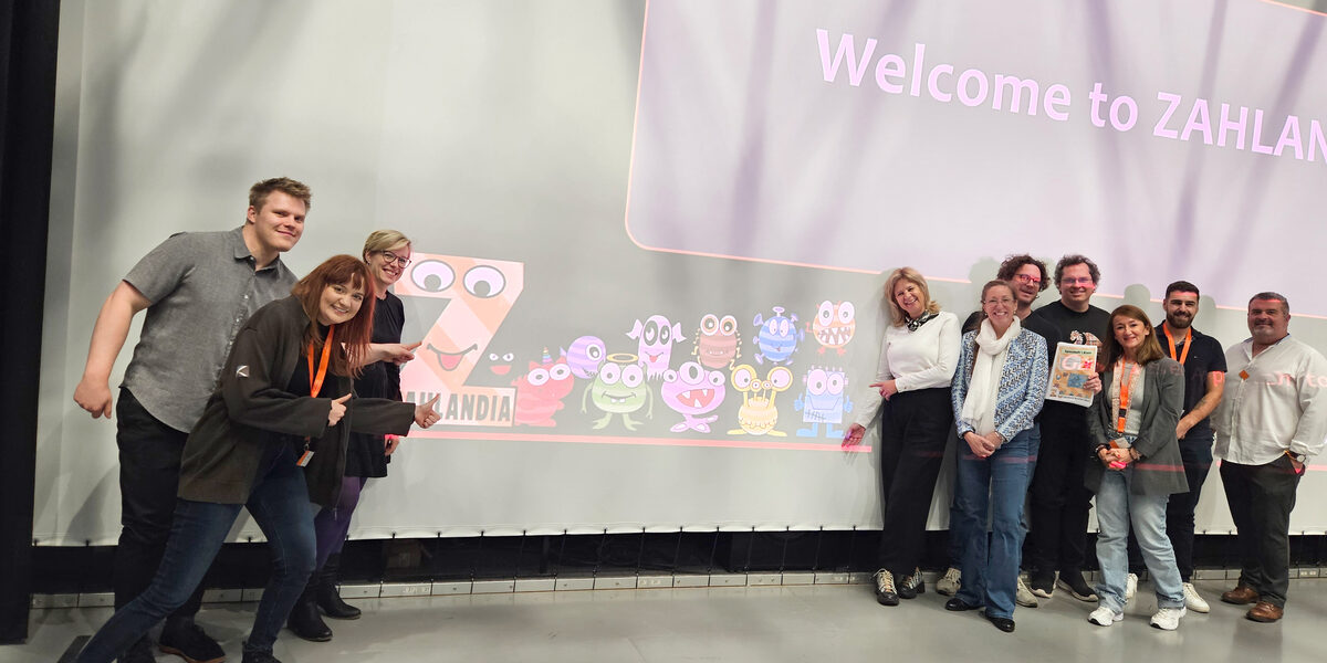 Zehn Personen stehen vor einer Leinwand, auf die "Welcome to ZAHLANDIA" und eine Reihe bunter Monsterfiguren projiziert ist.