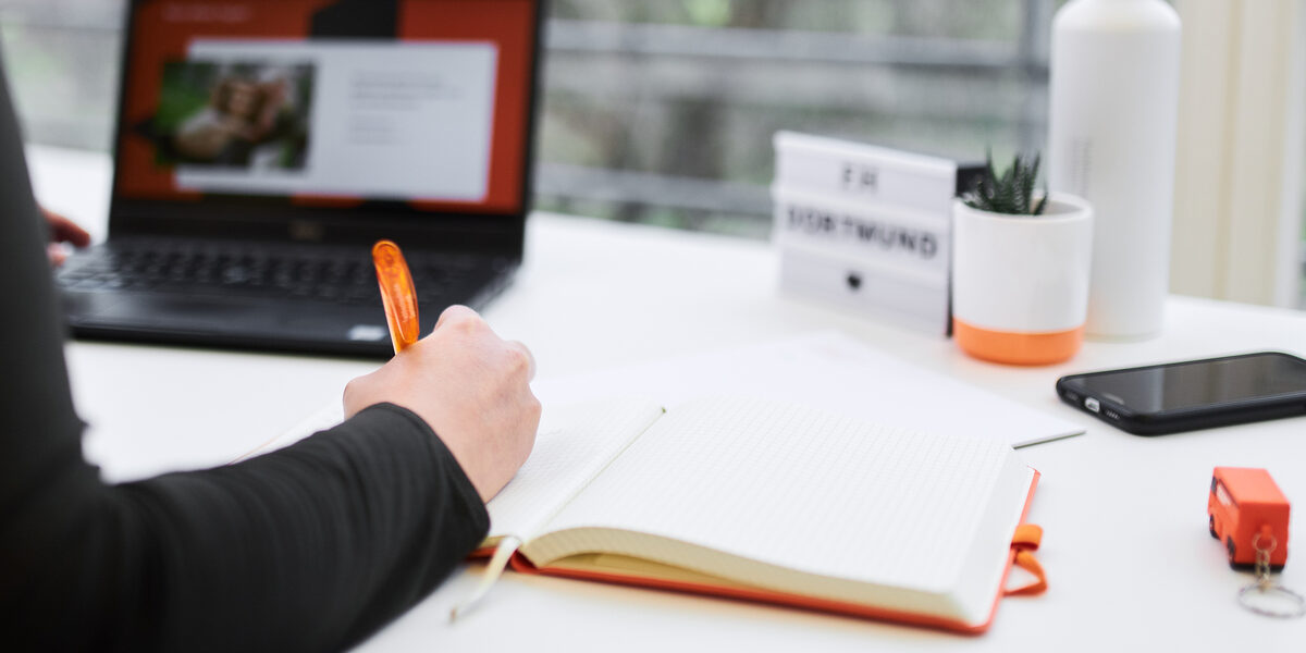 Foto eines Arbeitsplatzes mit Laptop. Im Vordergrund ist der Arm einer Person zu sehen, die etwas in ein Notizbuch schreibt.