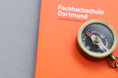 Photo of a small compass with a chain on it, lying with a pencil on an orange folder of the Fachhochschule Dortmund.