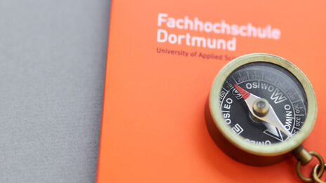 Foto eines kleinen Kompasses mit Kette daran, der mit einem Bleistift auf einer orangefarbenen Mappe der Fachhochschule Dortmund liegt.