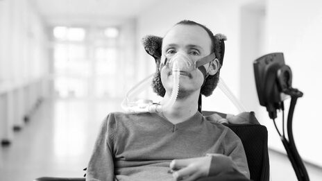 A person in a wheelchair with a breathing mask looks into the camera with a gentle smile.
