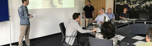 Study on quality of life. People sit and stand around a group of tables and look ahead at a presentation.