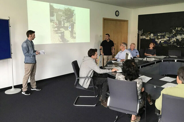 Studie zu Lebensqualität. Personen sitzen und stehen um eine Tischgruppe und blicken nach vorne auf eine Präsentation.