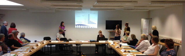 Conference on ageing and care. People sit and stand around a group of tables and look ahead at a presentation.