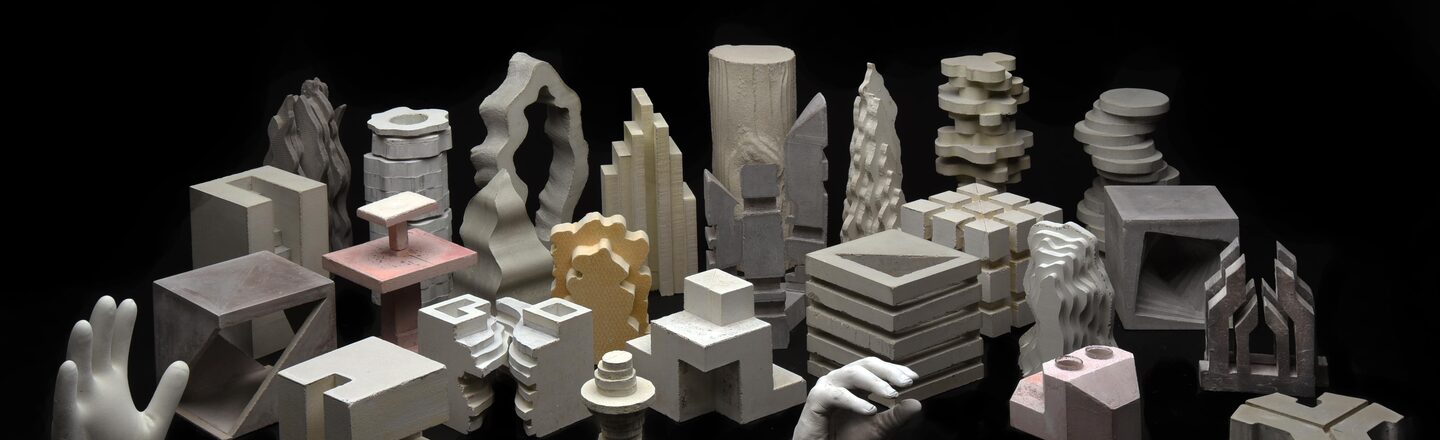 The picture has a narrow horizontal page format. It shows a collection of twenty-seven differently sized, dimensioned and generally differently designed sculptures made of concrete against a black background. The sculptures stand in approximately four rows and are offset in space. Their design appears geometric, free-form or a combination of both. Some of them are colored bright red, grey, black or yellow. At the back are tall, cylindrical or bar-shaped sculptures in terms of volume, but with very individual, sometimes curved and stepped shapes.