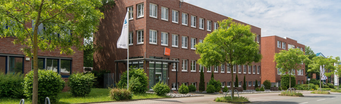 Photo of the Fachhochschule Dortmund site in Otto-Hahn-Straße from the outside. It is a red brick building. In the foreground are a road and some trees.