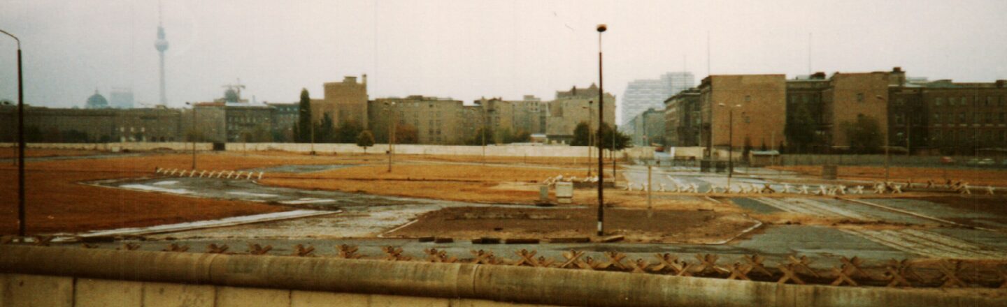 A picture of the Wall before it fell. You can see a wall covered in graffiti and a strip of empty space. There are buildings in the background.