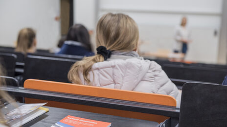 Foto einer jungen Frau in Rückansicht in einer Hörsaalreihe, diet nach vorne zur vortragenden Person schaut. In der Reihe dahinter ist der Tisch aufgeklappt, darauf liegen Printmaterialien. __ <br>Young woman in a row of lecture halls, looks forward to the speaker. In the row behind it, the table has been opened, and there are print materials on it.